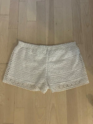 Vita spetsmönstrade shorts - Säljer ett par vita shorts med vackert spetsmönster och elastisk midja. Shortsen har en feminin look med fina detaljer längs bensluten och är perfekta för varma dagar. Passar dig som gillar en ljus och somrig stil.