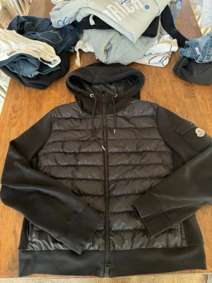 Svart Moncler Maglia zip cardigan - ✅ÄKTA✅Snygg svart zip-up cardigan från Moncler med vadderad front och mjuka ärmar. Jackan har huva med dragsko, två sidofickor med dragkedja och Moncler-logga på ärmen. Perfekt för lager-på-lager och streetwear-vibe.