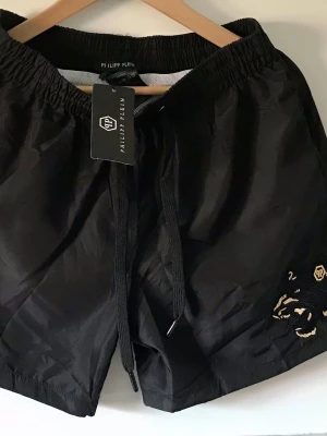 Svarta shorts/badbyxor från Philipp Plein - Svarta shorts/badbyxor från Philipp Plein med snörning i midjan och guldfärgad broderad detalj på ena benet. Materialet är syntet och de har en loose passform, perfekt för en avslappnad stil. Logotypen syns både på etiketten och broderiet.