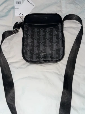Svart axelväska från Lacoste - Snygg svart axelväska från Lacoste med justerbar axelrem och geometriskt mönster i grått. Väskan har en fyrkantig form och är tillverkad i ett tåligt material som liknar konstläder. Perfekt för att bära det viktigaste och ger en stilren vibe.