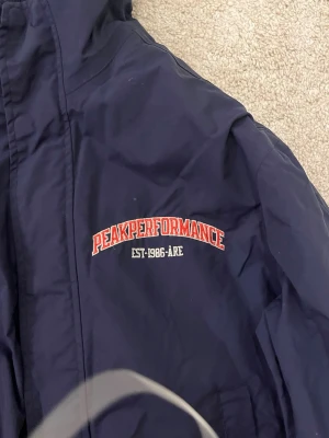 Marinblå vindjacka Peak Performance - Snygg marinblå vindjacka från Peak Performance med röd broderad logga på bröstet och siffran 86 på ärmen. Jackan har hög krage, dragkedja framtill och muddar vid ärmslut och midja. Perfekt för blåsiga dagar och sportig stil.