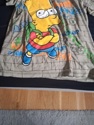 Beige t-shirt med Bart Simpson - Cool beige t-shirt med stort tryck av Bart Simpson på framsidan och graffitiliknande text i flera färger. T-shirten har ett rutigt mönster som liknar tegelvägg och är kortärmad. Perfekt för dig som gillar The Simpsons och streetstyle.