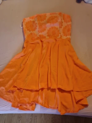Orange axelbandslös klänning från Zack - Snygg orange axelbandslös klänning från Zack med blommigt mönster upptill och vid kjol. Klänningen har en kort längd och är tillverkad i bomull, perfekt för varma dagar. Den har en flowy passform och är riktigt färgstark. Helt ny med prislapp, priset kan diskuteras
