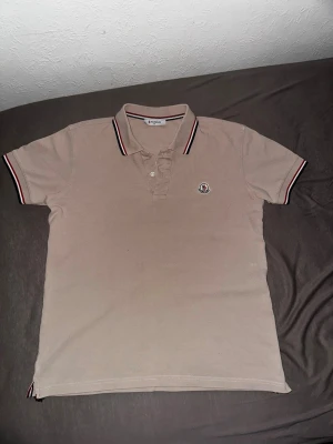 Beige pikétröja från Moncler - Snygg beige pikétröja med klassisk krage och knäppning framtill. Tröjan har korta ärmar och logga på bröstet. Kragen och ärmsluten har ränder i svart, vitt och rött för en sportig look.