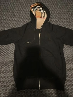 Svart Burberry hoodie med dragkedja - Svart hoodie från Burberry med klassiskt rutigt foder i huvan. Dragkedja framtill, fickor på sidorna och justerbar huva med snören. Ikonisk Burberry-logga broderad på bröstet. Perfekt för en stilren och trendig look.