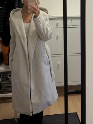 Ljusgrå lång kappa med huva - Snygg och stilren ljusgrå kappa med stor huva och öppen front. Kappan har en rak passform och är knälång, perfekt för lager-på-lager. Mjukt material som känns skönt mot huden. Snygg detalj med breda slag och diskreta fickor i sidorna.