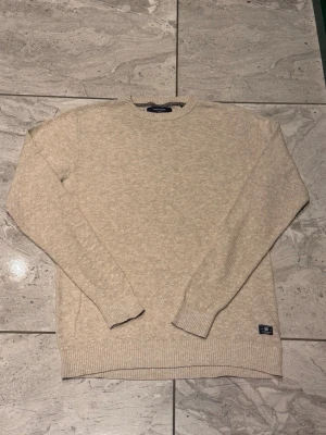 Beige stickad tröja från Jack and Jones  - Snygg beige stickad tröja från Jack and Jones med rund halsringning och ribbade muddar vid ärmslut och nederkant. Tröjan har en diskret logotyp-patch nertill. Mjuk och skön känsla i materialet.