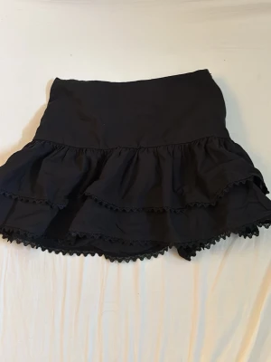 Svart volangkjol från Zara - Supersöt svart kortkjol från Zara med dubbla volanger och spetsdetaljer längst ner. Kjolen har en hög midja och är perfekt för att skapa en trendig och lekfull look. Tillverkad i mjukt material som känns bekvämt hela dagen.