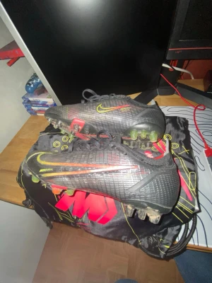 Nike Mercurial vapor 14 elite ag - Säljer ett par Nike Mercurial fotbollsskor i svart med neonfärgade detaljer i gult och rött. Skorna har en modern design med plastmaterial och mönstrad yta, samt dobbar för spel på konstgräs. Medföljer en snygg svart Mercurial-påse med grafiskt tryck.