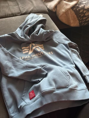 Blå hoodie från Alpha Industries - Snygg ljusblå hoodie från Alpha Industries med stor logga och text på bröstet. Hoodien har en klassisk känguruficka och ribbade muddar. Mjuk insida i fleece och huva för extra komfort. Perfekt för en avslappnad och cool stil.
