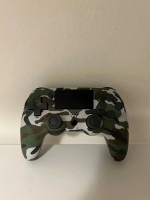 Kamo PlayStation 4 handkontroll - Säljer en PlayStation 4 handkontroll med snyggt kamouflagemönster i grönt, vitt, svart och brunt. Kontrollen har klassisk form med två analoga spakar, styrkors och touchpad. Tillverkad i plast och passar perfekt för gaming.