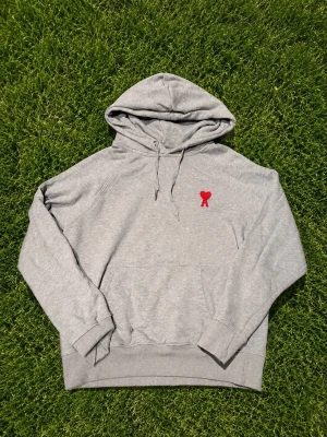 Grå hoodie från Ami Paris med röd logga - Snygg grå hoodie från Ami Paris med klassisk huva och dragsko. På bröstet sitter Ami Paris ikoniska röda hjärta med bokstaven A. Hoodien har känguruficka och ribbade muddar. Perfekt för en chill och stilren look.
