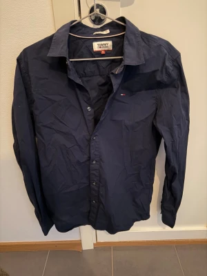 Mörkblå slim fit skjorta Tommy Jeans - Säljer en mörkblå skjorta från Tommy Jeans i slim fit-modell. Skjortan har klassisk krage, knäppning framtill och en liten logotyp på bröstet. Materialet är bomull och skjortan har långa ärmar. Perfekt för dig som gillar stilrena plagg med diskret branding.