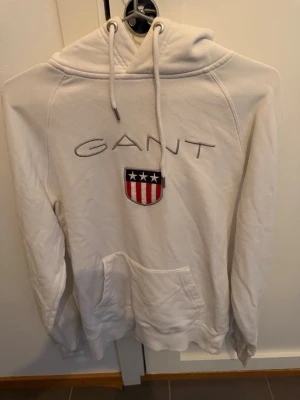 Vit hoodie från GANT med logga - Vit hoodie från GANT med broderad logotyp och amerikanskt inspirerat emblem på bröstet. Klassisk huva med dragsko och stor magficka framtill. Mjukt material och ribbade muddar vid ärmslut och nederkant. Perfekt för en avslappnad och cool look.