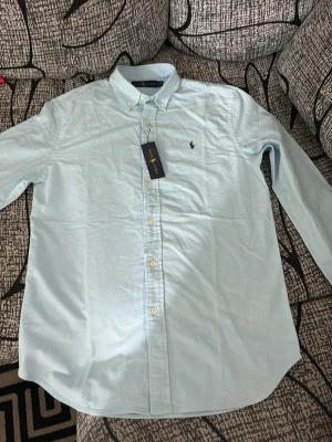 Ljusblå skjorta från Ralph Lauren - Klassisk ljusblå skjorta från Ralph Lauren med button-down krage och broderad logga på bröstet. Skjortan har långa ärmar och vita knappar framtill. Perfekt för en clean och stilren look.