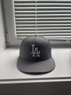 Grå LA-keps från New Era - Snygg grå keps från New Era med LA-logga broderad framtill i vitt. Klassisk flat peak och strukturerad form som ger en clean look. Perfekt för dig som gillar streetwear och sportig stil.