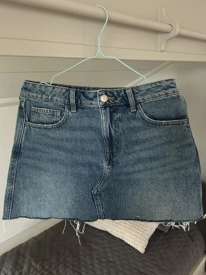 Blå jeanskjol med rå kant - Snygg blå jeanskjol i klassisk modell med fem fickor och rå, fransig kant nedtill. Kjolen har bälteshällor och stängs med knapp och dragkedja framtill. Perfekt för dig som gillar en avslappnad och trendig stil.