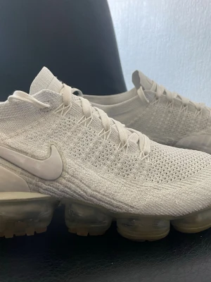 Nike Air Vapormax Flyknit vita/grå sneakers - Nike Air Vapormax Flyknit sneakers i helvitt och grå med stickad ovandel och genomskinlig sula. Skorna har en sportig look med diskret Nike-logga på sidan och elastiska skosnören. Ovandelen är i ventilerande textilmaterial och sulan har synliga luftbubblor för extra komfort. Storlek 44.5 för bara 389 kr