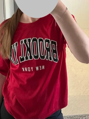 Röd Brooklyn New York t-shirt - Cool röd t-shirt med svart och vit text 'BROOKLYN NEW YORK' framtill. Klassisk passform med rund hals och korta ärmar. Perfekt för en avslappnad streetstyle-look.