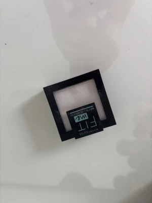 Maybelline Fit Me Matte + Poreless puder - Maybelline Fit Me Matte + Poreless foundationpuder i en fyrkantig svart plastförpackning med spegel. Färgen är ljus, nästan vit, och pudret ger en matt finish. Perfekt för dig som vill ha en naturlig look och kontrollera glans.