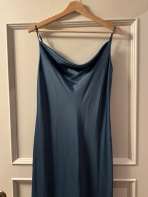 Blå långklänning i satin från MaxMara - Elegant blå långklänning från MaxMara i glansigt satinmaterial. Klänningen har smala axelband och en mjukt draperad urringning. Perfekt för dig som gillar en stilren och tidlös look.