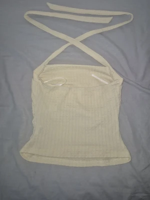 Beige ribbad linnetopp med knytband - Trendig beige linnetopp med ribbad struktur och smala knytband som korsas i nacken. Toppen är ärmlös och croppad, perfekt för en avslappnad och stilren look.