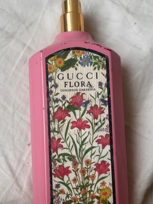 Gucci Flora Gorgeous Gardenia parfym - Gucci Flora Gorgeous Gardenia parfym i en lyxig rosa flaska med guldfärgad spray och blommigt  mönster på etiketten. Flaskan har en elegant, kantig form och detaljer i guld. Perfekt för dig som gillar exklusiva och feminina dofter. Den har ungefär lite mer än hälften kvar, och hela flaskan är rätt skrapad och har förlorat färg på ställen, men sprayen funkar precis som den ska.