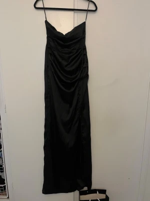 Svart långklänning med slits - Elegant svart långklänning med tunna axelband och draperad framsida. Klänningen har en snygg slits på sidan och är tillverkad i ett glansigt material som ger en lyxig känsla. Perfekt för dig som vill sticka ut med en stilren look.