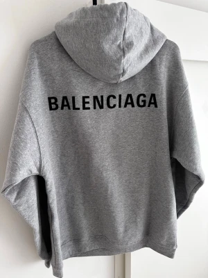 Grå Balenciaga hoodie helt ny med tags - Storlek xs men passar större S/mindreM Snygg grå hoodie från Balenciaga med svart logotyptryck på bröstet och stort på ryggen. Klassisk känguruficka framtill, dragsko i huvan och mjukt material. Perfekt för en avslappnad streetwear-look.