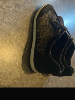 Bruna Fendi sneakers med monogram - Snygga bruna sneakers från Fendi med klassiskt FF-monogram på sidorna. Skorna har mockadetaljer, bruna skosnören och vit sula. Perfekta för dig som gillar lyxiga och trendiga märken med unik design.