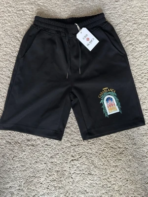 Svarta shorts från Casablanca - Svarta shorts från Casablanca med färgglad tryckt grafik på ena benet och texten 'Avenida Casablanca'. Shortsen har elastisk midja med snörning och sidofickor. Perfekta för sommaren och har en avslappnad passform.