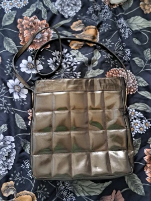 Unik Vintage Charles Jourdan väska – Made in Iran – Quiltat läder - Säljer en mycket elegant axelremsväska från det franska lyxmärket Charles Jourdan. Detta är ett ovanligt samlarobjekt då den är märkt **"Made in Iran"** på insidan. Denna märkning härrör sannolikt från märkets licenstillverkning före 1979, vilket gör den till en unik del av modehistorien.  Design: Klassiskt quiltat mönster med en sofistikerad brons/metallic-finish. Material: Läder med metallic-yta, insida i brunt läder/foder. Detaljer: Dragkedja upptill samt ett mindre innerfack med dragkedja.