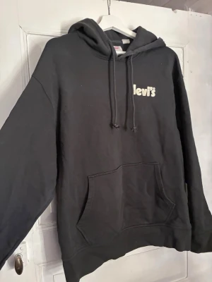 Svart hoodie från Levi's med logga - Svart hoodie från Levi's med vit logga på bröstet. Klassisk modell med huva och dragsnören samt stor känguruficka framtill. Mjuk bomullsblandning som är perfekt för en avslappnad streetstyle-look.
