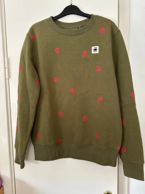 Olivgrön sweatshirt från G-Star RAW, storlek S man  S - Cool olivgrön sweatshirt från G-Star RAW med röd/orange tryckdetaljer över hela tröjan. Rund halsringning och ribbade muddar vid ärmslut och nederkant. Perfekt för en avslappnad och trendig stil. Supermjuk å skön. Använd ett par gånger. 