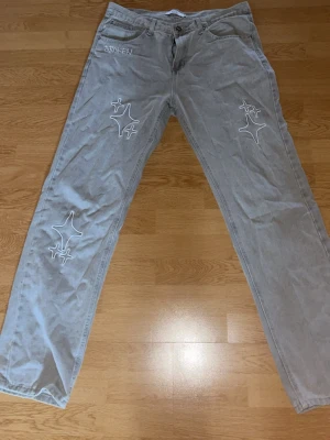 Grå jeans Broken Planet Market - Säljer ett par grå jeans från Broken Planet Market med vita broderade stjärndetaljer på benen och texten 'BROKEN' vid fickan. Jeansen har rak passform, fem fickor och är gjorda i denim. Perfekta för dig som gillar streetwear och unika detaljer.