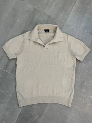 Beige stickad polotopp Massimo Dutti - Snygg beige stickad polotopp från Massimo Dutti. Toppen har korta ärmar, bred krage och ribbade detaljer vid ärmslut och nederkant. Perfekt för en stilren och avslappnad look.