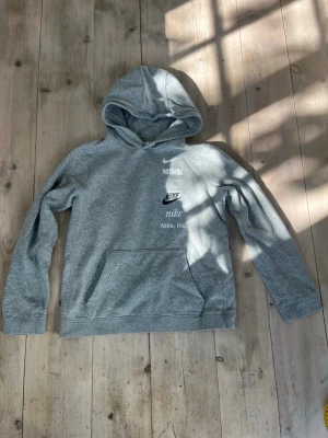 Grå hoodie från Nike med loggor - Snygg grå hoodie från Nike med flera olika Nike-loggor tryckta på bröstet i vitt och svart. Hoodien har en klassisk huva och en stor magficka framtill. Tillverkad i mjukt sweatshirtmaterial som är skönt att ha på sig. Perfekt för en avslappnad och sportig stil. Stl passande dig som är 158-164 cm lång.
