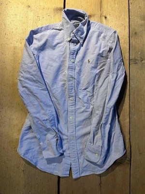 Ljusblå skjorta från Polo Ralph Lauren - Klassisk ljusblå skjorta från Polo Ralph Lauren med broderad logga på bröstet. Skjortan har lång ärm, knappar framtill och button-down krage. Modellen är custom fit och passar perfekt till en stilren look.
