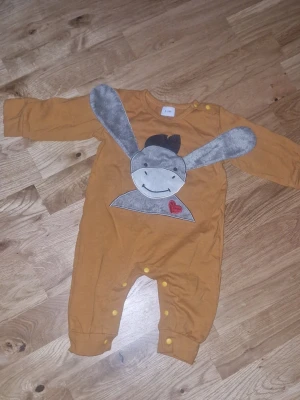 Bebis overall unisex - Söt gul sparkdräkt med lång ärm och tryckknappar längs benen. Framsidan har ett stort motiv av en glad åsna med mjuka, grå öron och ett rött hjärta. Tillverkad i mjuk bomull, perfekt för små barn. Storlek 2-4M. Inga defekter.