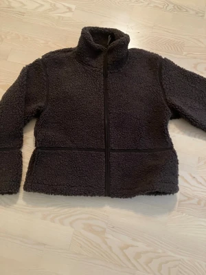 Mörkbrun teddyjacka ( Pilejacka)  med dragkedja - Mysig mörkbrun teddyjacka med hög krage och hel dragkedja framtill. Jackan har en boxig passform och är tillverkad i fluffigt teddy-material som ger en riktigt skön och trendig look. Perfekt för kyliga dagar när du vill vara både varm och snygg.