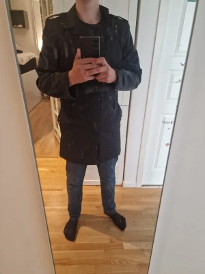 Svart trenchcoat med axelklaffar - Stilren svart trenchcoat med axelklaffar och knäppning framtill. Jackan har en rak passform och sidofickor med knappdetaljer. Perfekt för dig som vill ha en klassisk och tidlös look. Materialet ser ut att vara bomull eller en bomullsblandning. Pris kan diskuteras