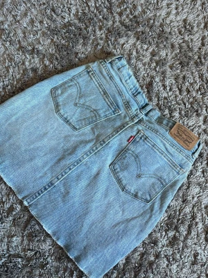 Levi's ljusblå jeanskjol - Snygga ljusblå jeansshorts från Levi's med klassisk femficksdesign, rå kant nertill och hög midja. Ikonisk läderpatch baktill och den röda Levi's-taggen på ena bakfickan. Perfekta för sommaren och enkel att matcha med allt.