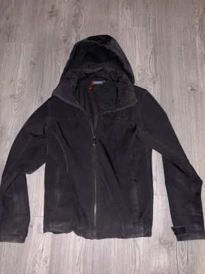 Svart vindjacka från Sail Raising - Svart vindjacka med huva från 8848 Altitude. Jackan har dragkedja framtill, två sidofickor och justerbar kardborre vid ärmslut. Materialet är lätt och vindtätt, perfekt för blåsiga dagar. Snygg och stilren design med diskret logga på ärmen.