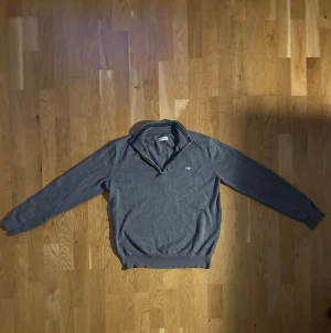Grå halvzip-tröja från Gant - Snygg grå tröja från Gant med halv dragkedja och broderad logga på bröstet. Tröjan har ribbade muddar och krage, perfekt för lager-på-lager-stil. Klassisk och stilren design som funkar till det mesta. Knappt använd