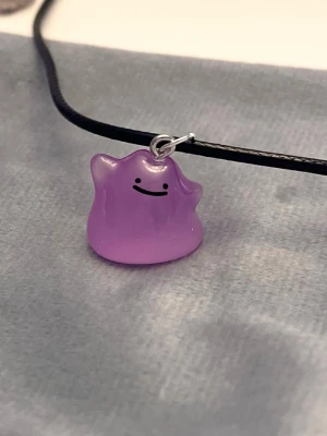 Självlysande Ditto halsband Pokémon smycke - Så gulligt halsband med en berlock av Ditto!Berlocken är ca 1,5 x 1,5 cm och trädd på svart tråd. Den är också självlysande och lyser i mörkret. Halsbandet är justerbart 40-45 cm. 