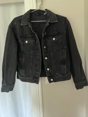 Svart jeansjacka  - Snygg svart jeansjacka med krage och silvriga knappar framtill. Jackan har två bröstfickor med lock och knapp samt markerade sömmar. 