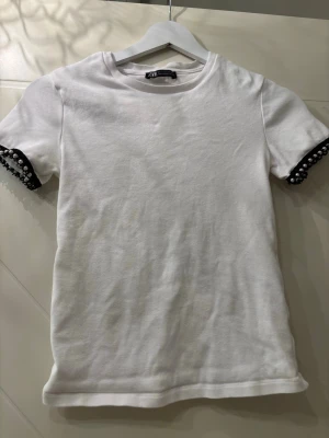 Vit t-shirt med pärldetaljer från ZARA - Vit t-shirt från ZARA med korta ärmar och rund halsringning. Ärmsluten är dekorerade med svarta pärldetaljer som ger en unik touch. Tillverkad i mjuk bomull för en skön känsla. Perfekt för dig som vill ha något stilrent men ändå speciellt.