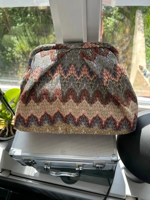 Mönstrad kuvertväska i tyg - Säljer en unik kuvertväska i tyg med färgglatt zigzag-mönster i beige, blått, rött och grönt. Väskan har en klassisk form och stängs upptill. Insidan är fodrad i ljusbrunt tyg och har en liten innerficka med dragkedja. Perfekt accessoar för att lyfta din outfit.