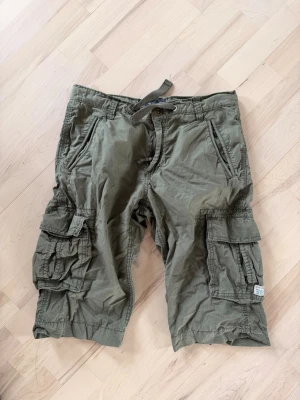 Olivgröna cargoshorts med snörning - Säljer ett par olivgröna cargoshorts med flera stora fickor på sidorna och baktill. Shortsen har snörning i midjan och är tillverkade i ett slitstarkt bomullsmaterial. Perfekta för dig som gillar en loose passform och praktiska detaljer.