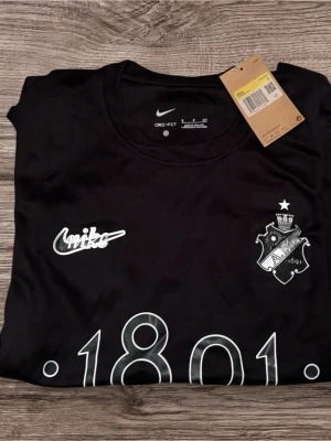 Svart AIK t-shirt från Nike - Svart t-shirt från Nike med AIK:s klubbmärke och texten '1891' tryckt på framsidan. Tröjan har rund halsringning och är tillverkad i Dri-FIT-material som andas, perfekt för träning eller match. Snygg och stilren design med vita detaljer.